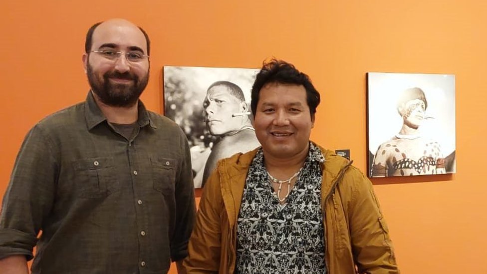 Fotografia colorida mostra dois homens, um branco e um indígena, lado a lado em frente a uma parede com fotografias