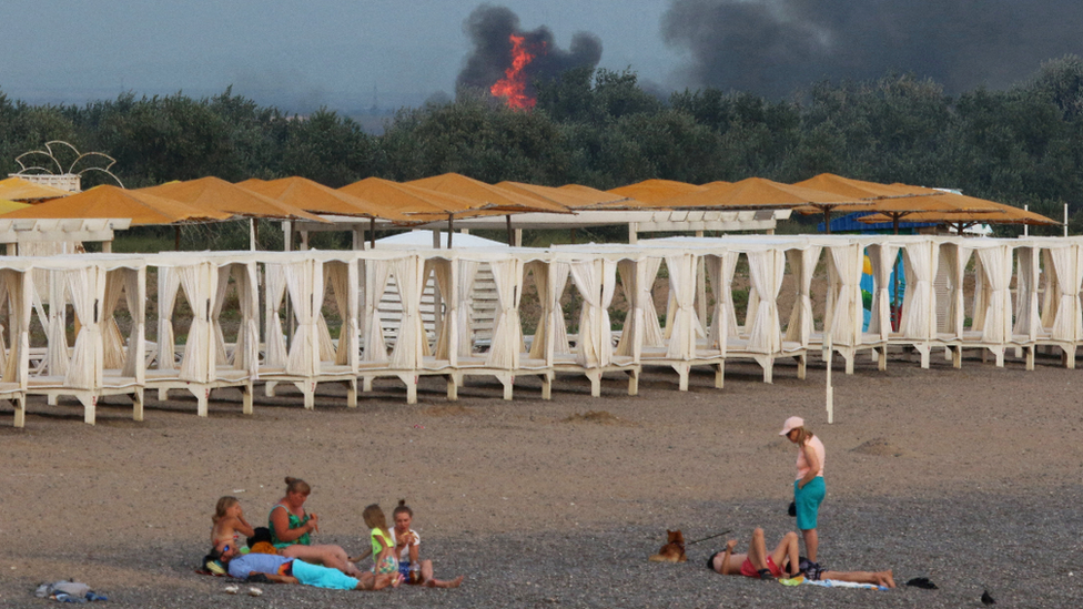 Turistas disfrutan de la playa mientras asciende una nube de humo en una base militar rusa