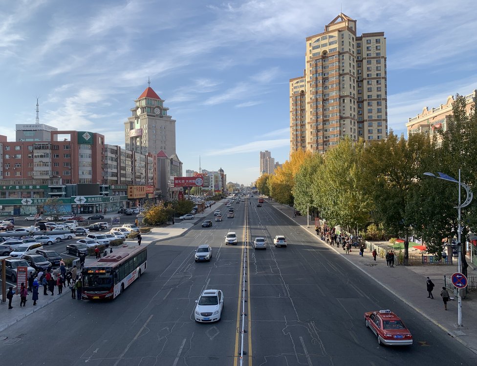 Городской пейзаж с автомобилями, едущими по прямой дороге, окруженной зданиями и автостоянкой