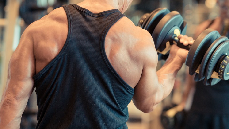 Muscular man lifting dumbbell with one hand on back in gym