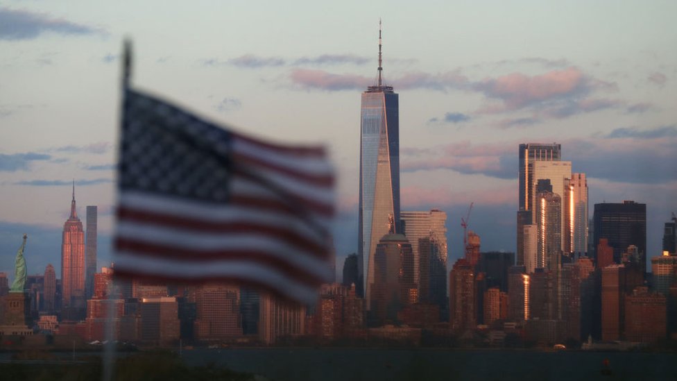 Американский флаг развевается перед Статуей Свободы, Эмпайр-стейт-билдинг и Всемирным торговым центром на закате в Нью-Йорке 15 декабря 2019 г., если смотреть из Байонны, штат Нью-Джерси