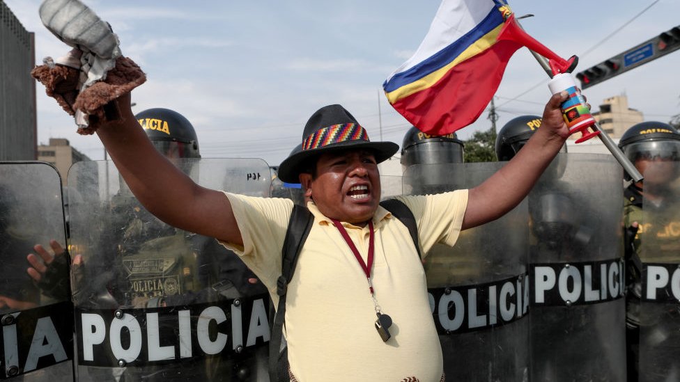 Un hombre protesta y enarbola unas banderas frente a un cordón policial en Perú.