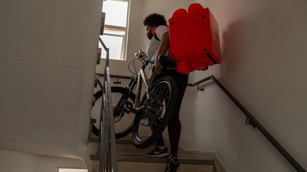 Franciele sobe escada carregando mochila de delivery e bicicleta
