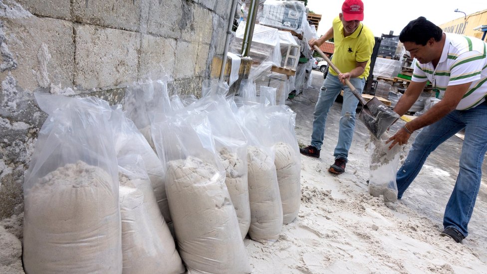 Los sacos de arena se usan para rodear las construcciones y así frenar el paso del agua. Un señor llena el saco de otro señor con arena, utilizada para frenar el paso del agua
