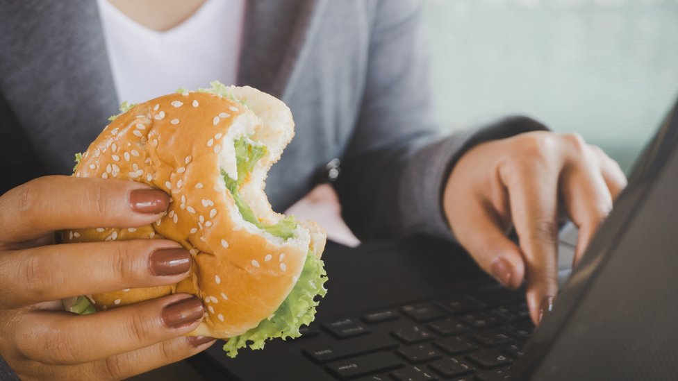 Una mujer frente a una computadora con una hamburguesa.