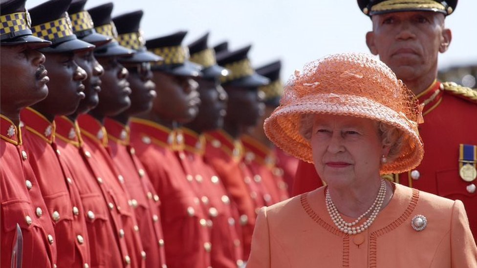 A rainha Elizabeth, uma senhora branca idosa de terno e chapéu rosas, passa em frente a vários homens negros fardados em uma visita à Jamaica em 2002