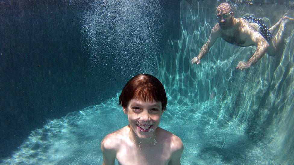 Un niño sonríe con un adulto de fondo debajo del agua