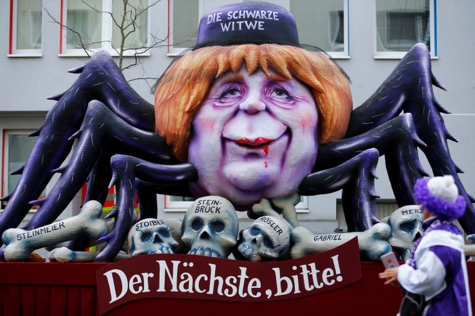 Lutka sa karnevala u Diseldorfu iz gebruara 2018. prikazuje nemačku kancelarku Angelu Merkel kao crnu udovicu.. Na transparentu piše: „Sledeći, molim!"