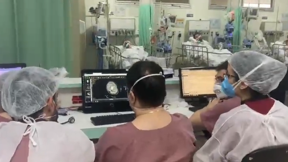 Médicos reunidos em torno do computador, com pacientes ao fundo