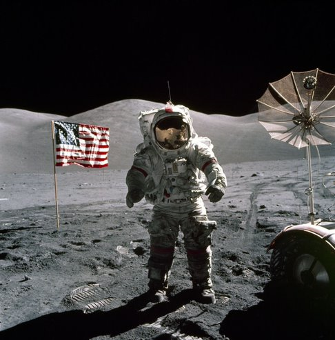Astronauta da Apollo 17 em pé sobre a superfície lunar com a bandeira dos Estados Unidos e uma antena parabólica ao fundo