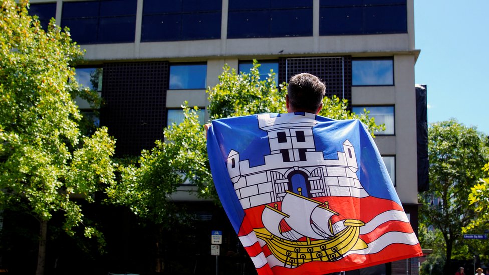 Homem envolto com a bandeira da Sérvia em frente a hotel