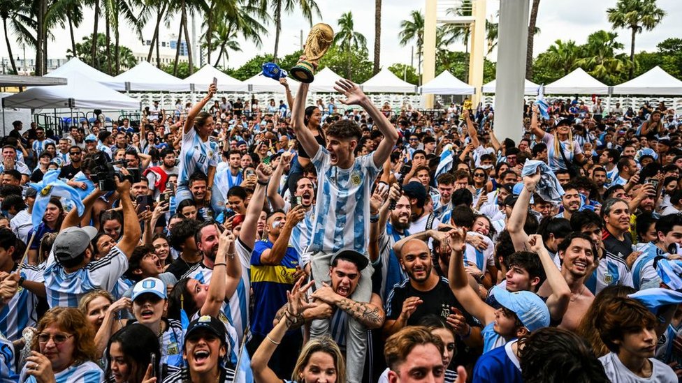 Personas celebrando el triungo de la selección. En el centro, un joven alza un trofeo réplica de la Copa del Mundo