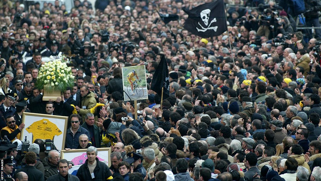 Marco Pantani's coffin is carried through a packed street, with winning jerseys, portraits and skull and crossbones flags held aloft