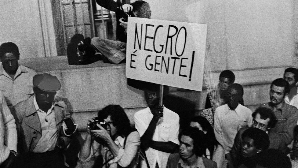 Manifestação do Movimento Negro Unificado, 1980. No centro da foto, manifestante com cartaz onde se lê: 'Negro é gente'