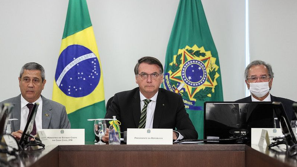Braga Netto, Bolsonaro e Guedes sentado em mesa de reunião, com feições sérias e bandeiras no plano de fundo