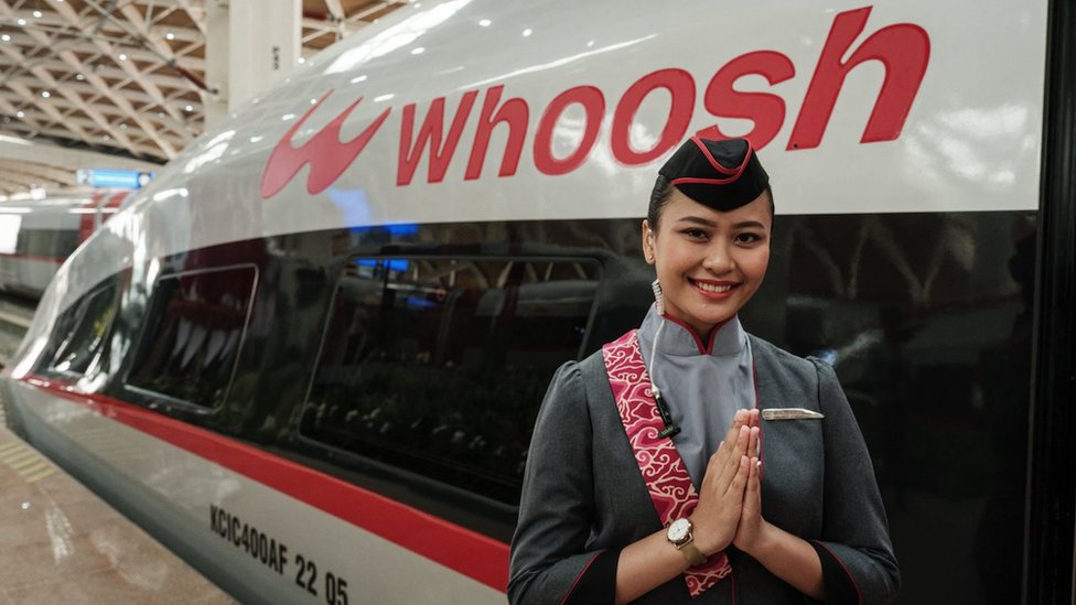 A Whoosh staff member holds her hands together in a welcoming gesture in front of the Whoosh train