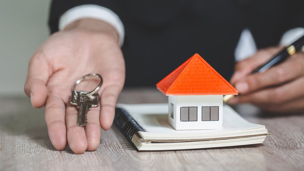 Un hombre firma un documento con una pequeña maqueta de una casa, mientras en la otra mano ofrece unas llaves