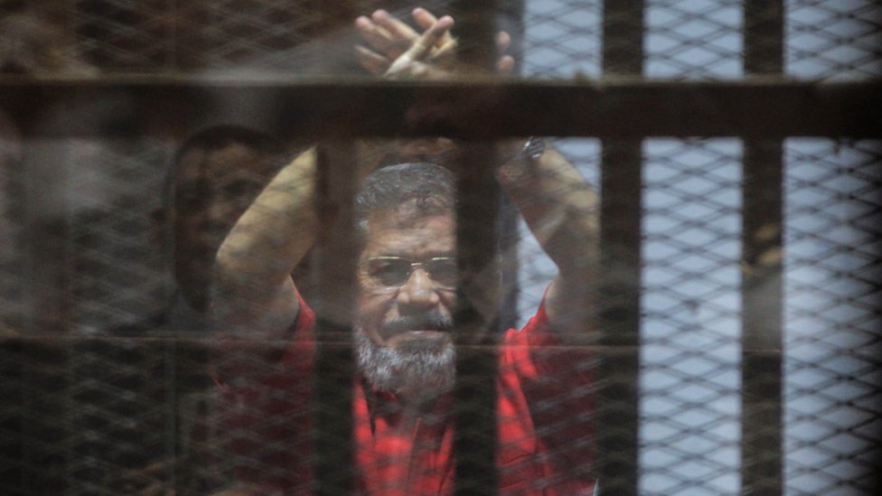 Бывший президент Египта Мохамед Мурси поднимает руки во время выступления в суде