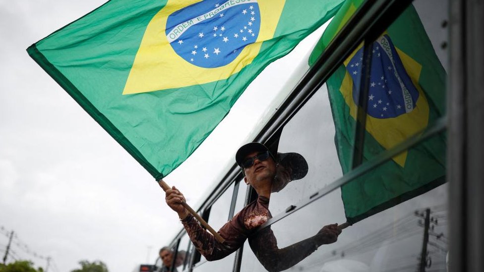 Un hombre sostiene la bandera de Brasil desde la ventana de un autobús al ser detenido luego de la invasión de Brasilia por seguidores de Bolsonaro.