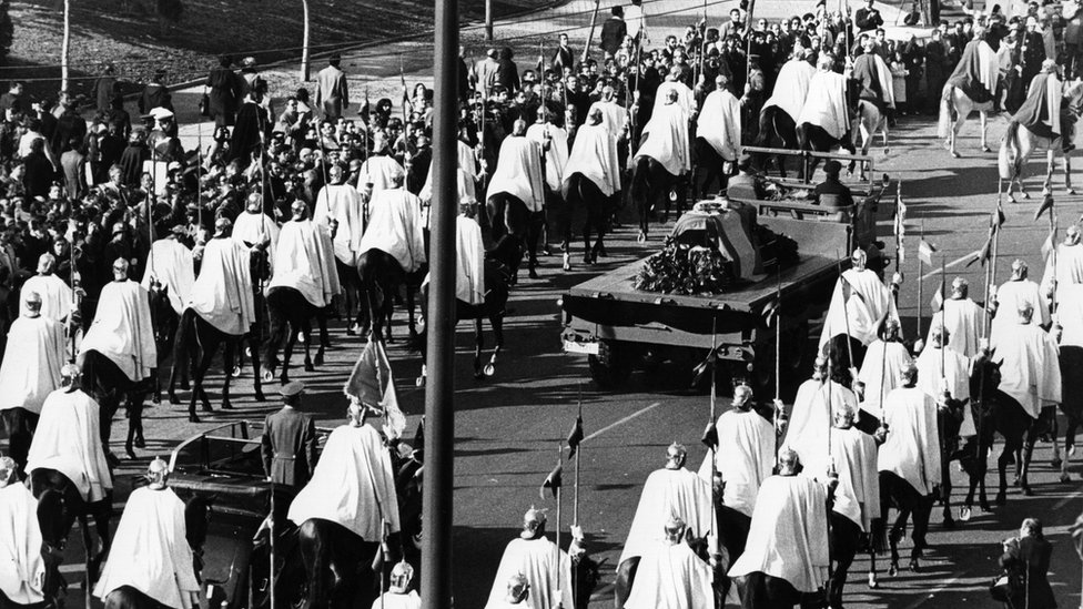 Похоронная процессия Франко в 1975 году в сторону Долины павших. Его гроб, задрапированный флагом, окружают люди в белых плащах на лошадях
