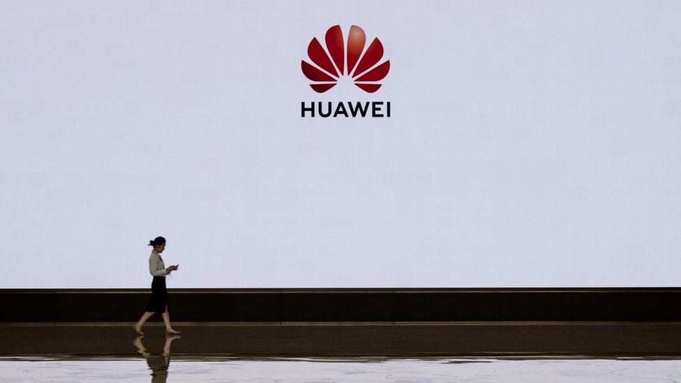 Сотрудник приемной Huawei идет перед большим экраном с логотипом в фойе здания, которое использовалось для посещений высокопоставленных клиентов и дисплеев в кампусе компании Bantian 12 апреля 2019 года в Шэньчжэне, Китай.
