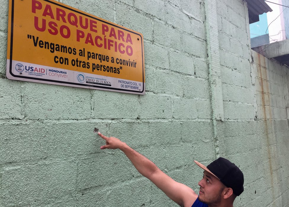 Kelvin señala un orificio de bala bajo un cartel de "Parque para uso pacífico".