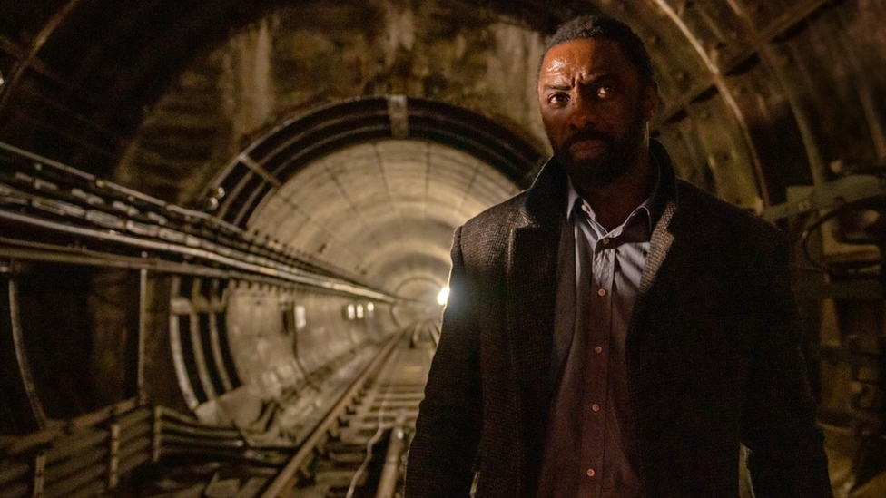Um homem negro de barba com expressão preocupada anda em um túnel mal iluminado