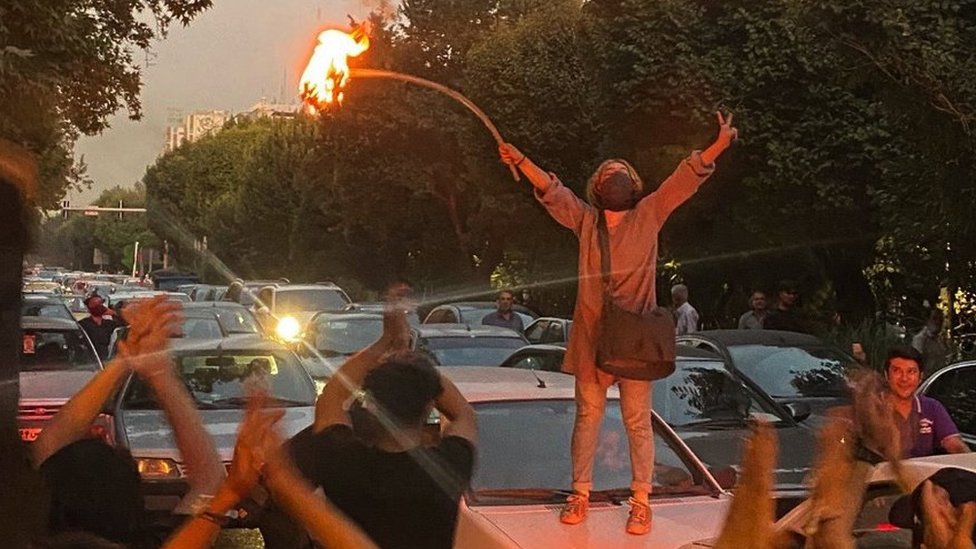 A woman stands on top of a car in the road, holding a stick on fire