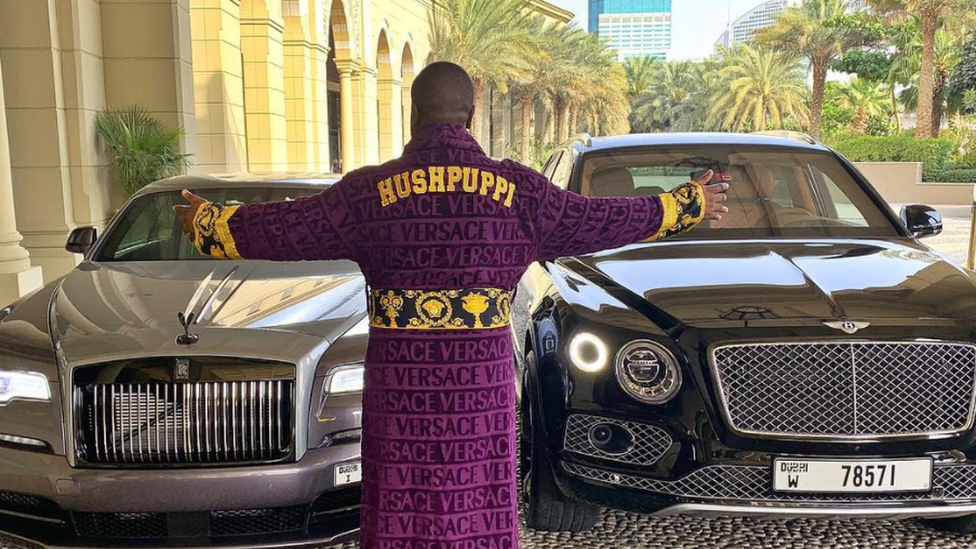 Hushpuppi in a monogrammed dressing gown by some luxurious cars