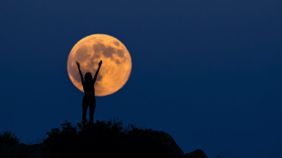 Una luna llena anaranjada, con una mujer frente a ella, con los brazos en alto.