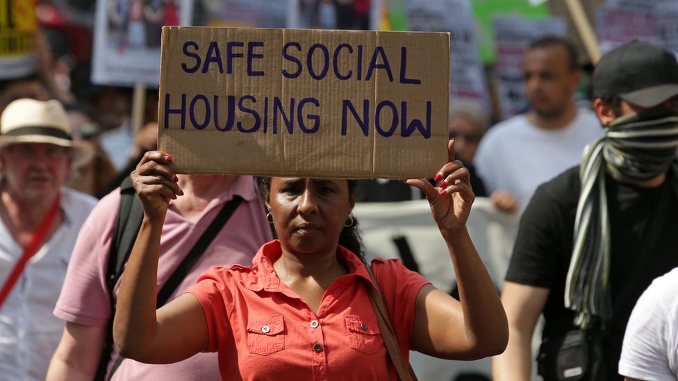 женщина держит плакат с надписью «теперь безопасное социальное жилье»