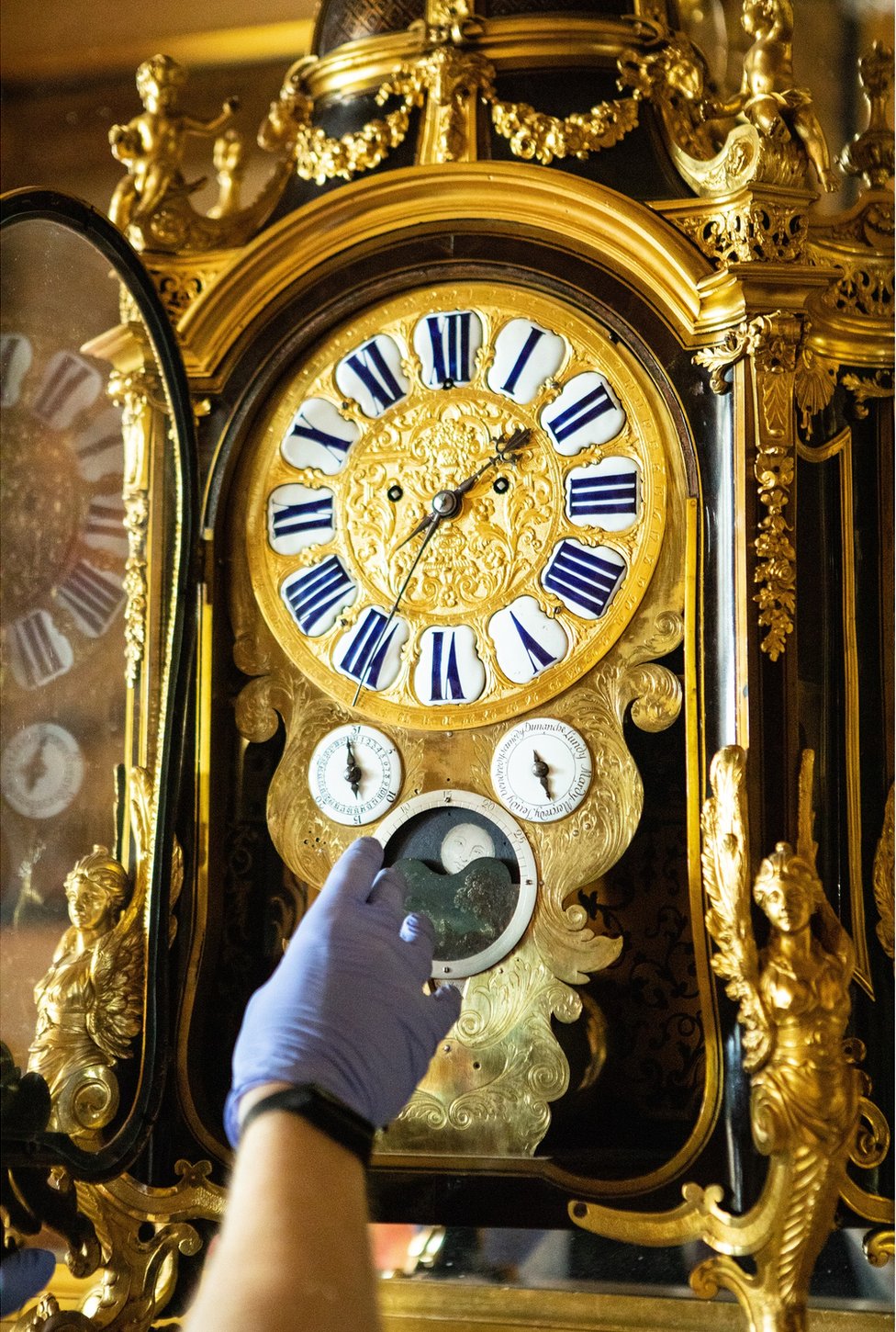 A close-up of Fjodor changing the time on a gold-decorated clock with a dial for moon changes