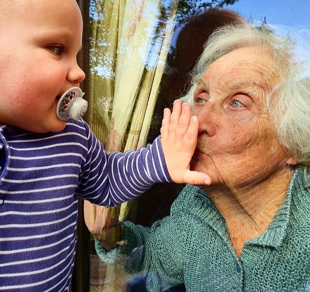 Young child and his great grandma greet each other through the window pane