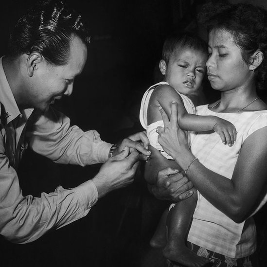 Um homem administra uma vacina a uma criança segurada por uma mulher
