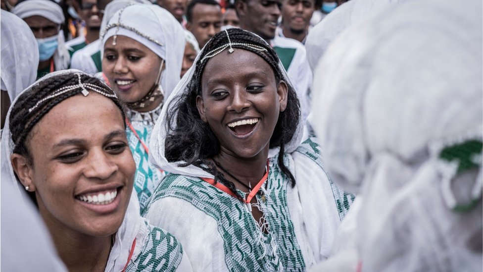 Smiling Ethiopian women dressed in traditional clothing celebrating the swearing in of Prime Minister Abiy Ahmed. They are part of a much larger crowd it seems. They are wearing intricate head accessories. They all look very happy.