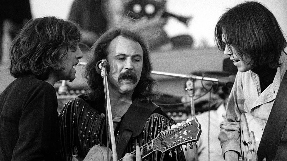 Graham Nash, David Crosby and Neil Young sing into a microphone during a concert in 1969