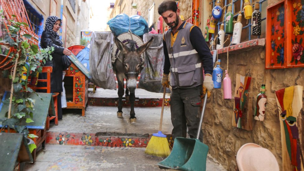 Un trabajador del aseo municipal limpia las calles y detrás lleva a un burro que carga las bolsas de basura.