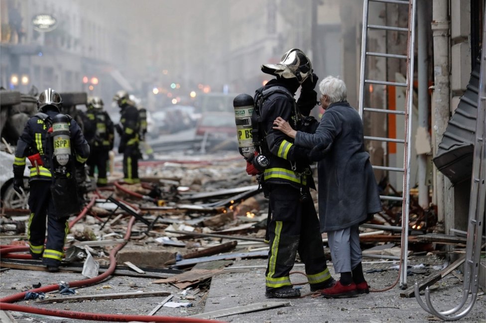 An elderly woman evacuated from a damaged building holds the arm of a fiefighter