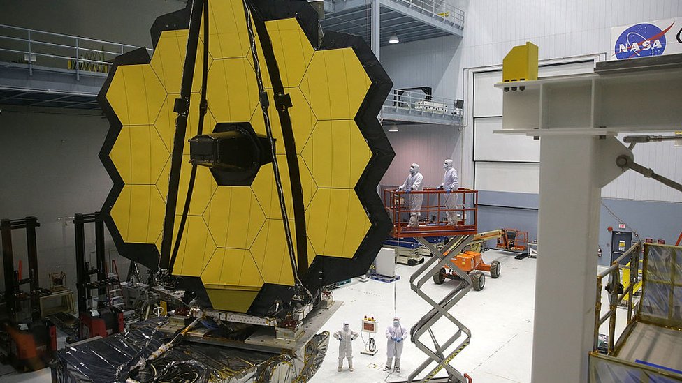 Telescópio James Webb em grande sala, com pessoas de jaleco trabalhando em volta dele