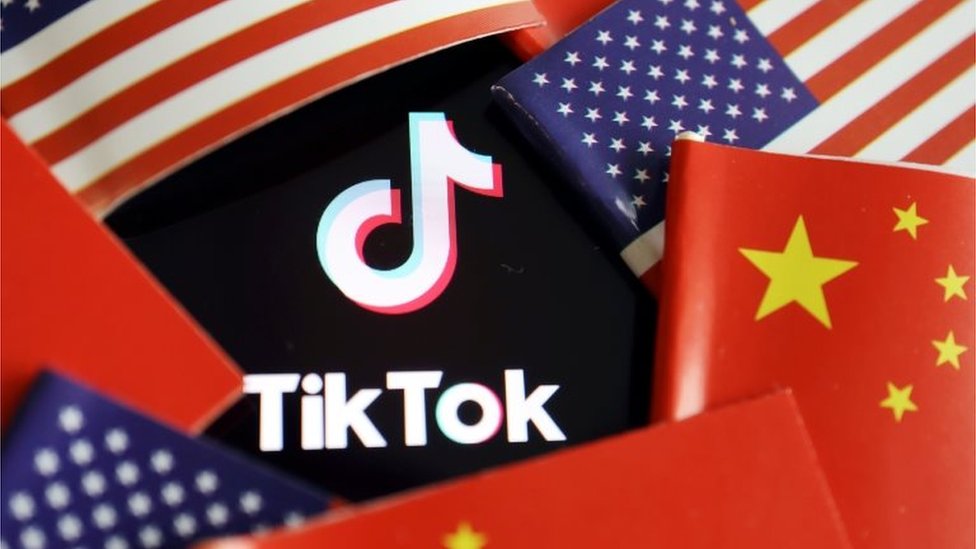 Здесь можно увидеть логотип TikTok, частично покрытый кольцом чередующихся флагов США и Китая