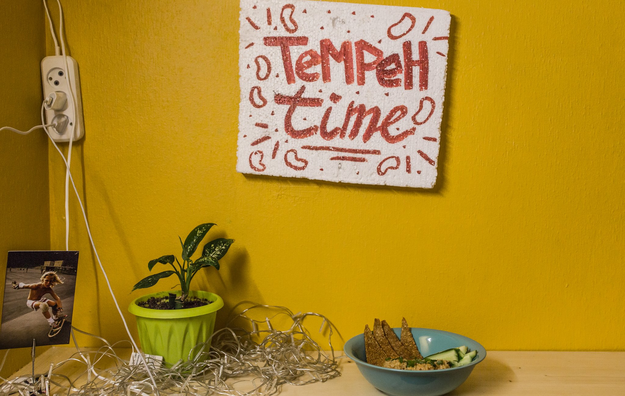 Polystyrene sign with 'Tempeh Time' painted on it in red above a bowl of food, on a bright yellow wall