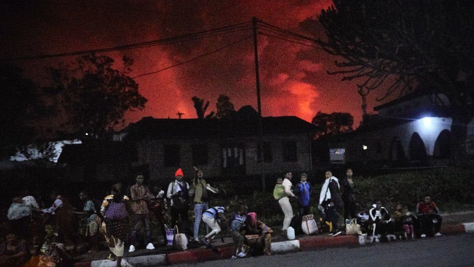 أشخاص منتظرون على رصيف ومن ورائهم السماء حمراء بفعل أدخنة البركان الثائر