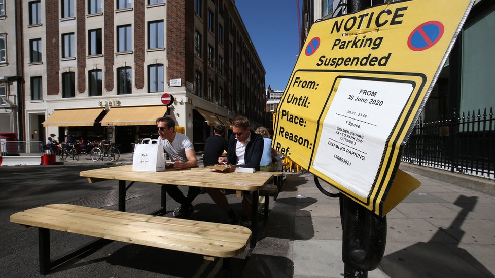 People eat lunch at tables and benches placed in the roadway adjacent to a suspended parking bay in Golden Square, Soho
