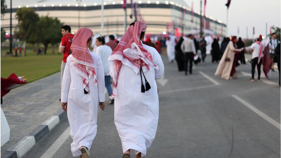 Dos hombres con túnica blanca, conocida como thaub, y pañuelo rojo y blanco -llamado kufiya- caminan hacia el estadio Al Bayt.
