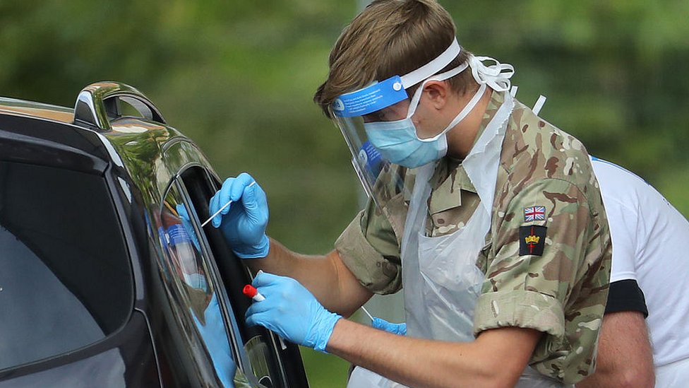 мужчина в военной форме в маске, перчатках и козырьке делает мазок через окно машины