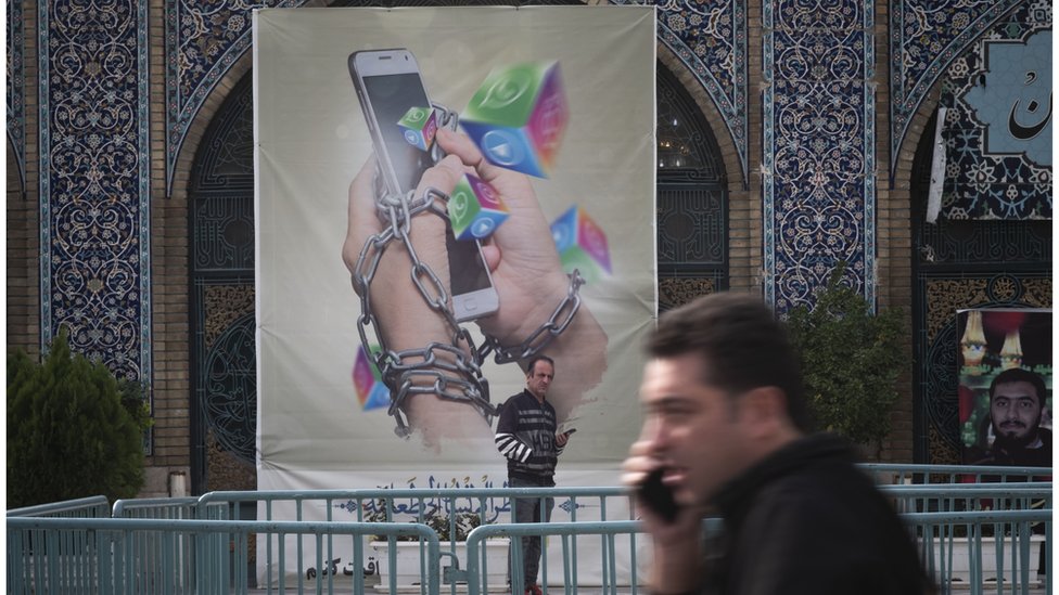 رجل إيراني يحمل هاتفه الذكي ينظر إليه وهو يقف تحت لافتة مناهضة لوسائل التواصل الاجتماعي في سوق طهران الكبير في 3 ديسمبر/ كانون الأول 2022.