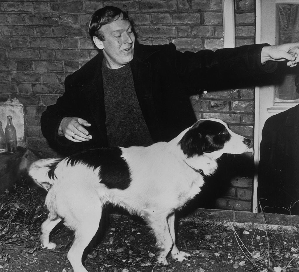 David Corbett, vestido con chaque, hace una señal a su perro con el brazo izquierdo y el perrito Pickles, en el centro de la imagen, mira hacia ese lado.
