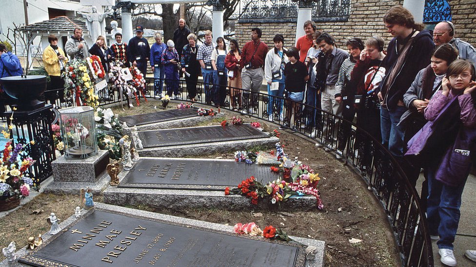 Fãs se aglomeram em torno da pequena cerca que os distancia do túmulo de Elvis, no centro, cercados por seus familiares