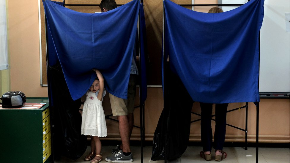 Ребенок смотрит из-за занавески кабины для голосования, пока взрослые голосуют на выборах в Греции