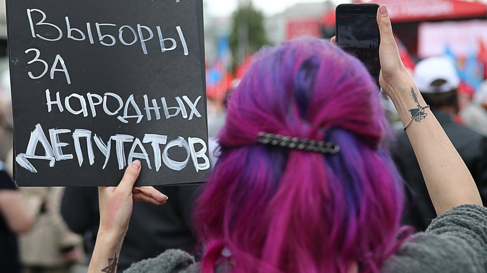 Женщина спиной к камере держит табличку с текстом на русском языке.